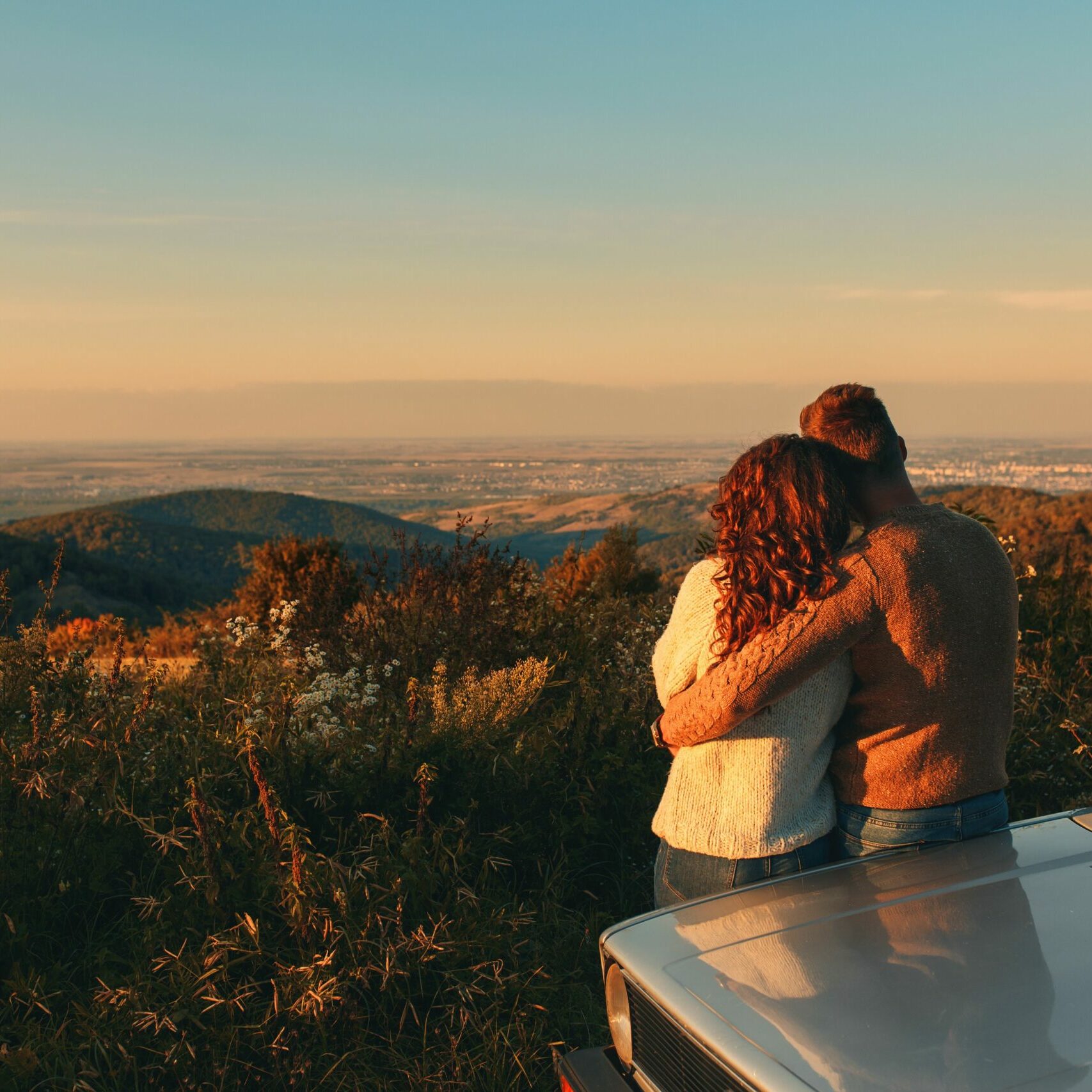 Rear view of beautiful young couple resting after road trip leaning against car and enjoying the view.